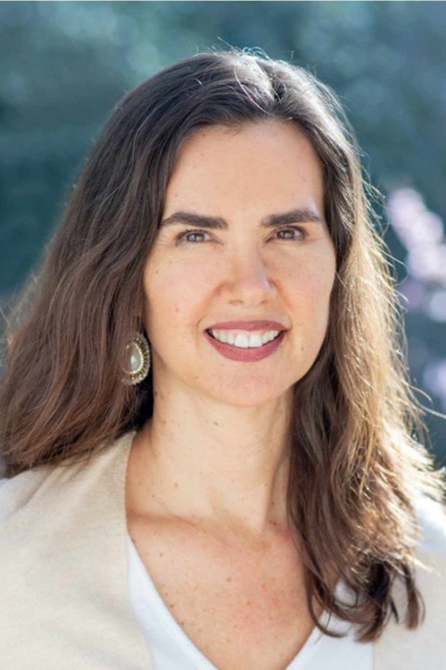 Dr. Kristin Neff smiling outdoors, with long brown hair, a light-colored top, and a beaded earring, softly lit against a blurred natural background.